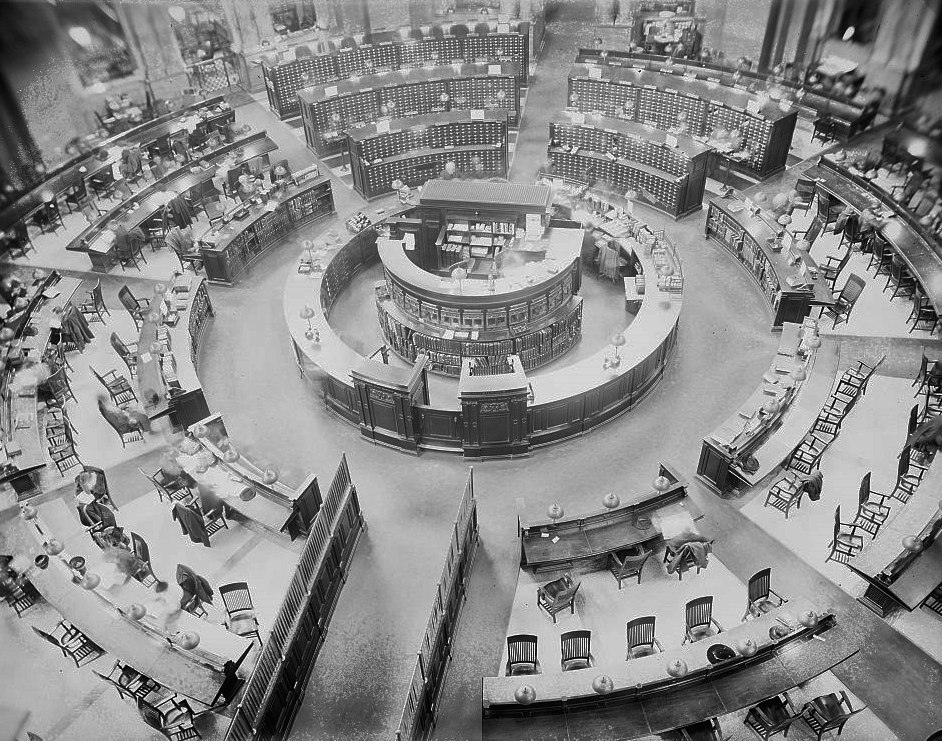 A bird's-eye view of the circular reference desk at the center of the Library of Congress Main Reading Room, ringed by card catalogs and reading chairs radiating outward