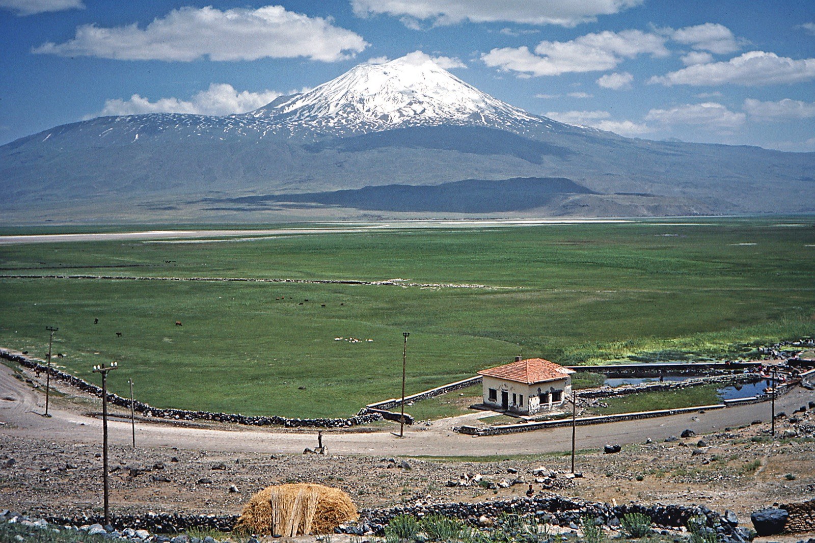 Snow-capped Mount Ararat rising over a wide green plain in eastern Turkey, with a small stone building and a hay bale in the foreground