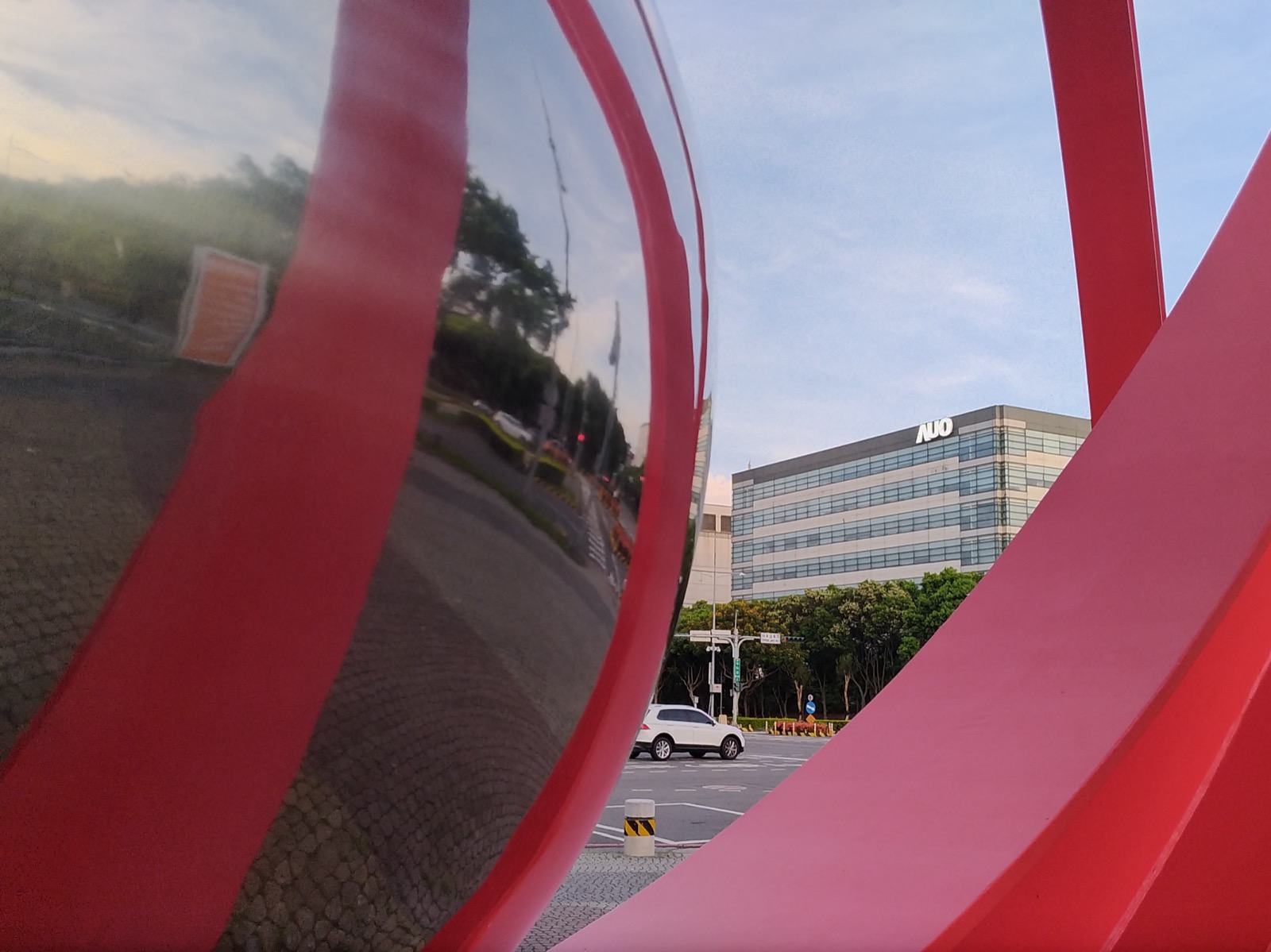 An AUO office building in Taichung's Central Taiwan Science Park at dusk, its blue-green glass curtain wall and white AUO logo seen through the gap in a large red public sculpture whose polished mirrored inner surface reflects the road, trees and passing traffic