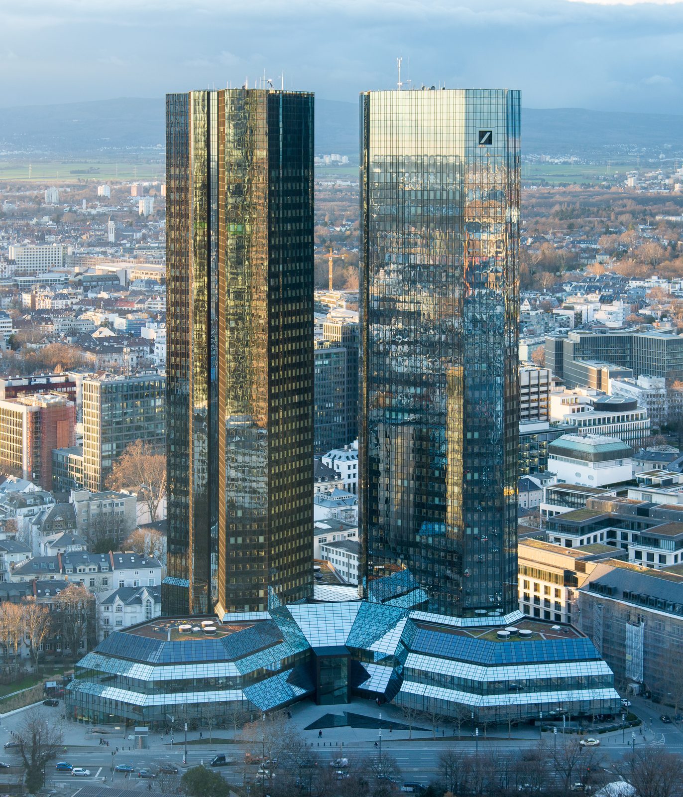 The mirrored glass Twin Towers of Deutsche Bank's Frankfurt headquarters, photographed from the southeast in warm evening light, the bank's stylised slash-in-a-square logo visible near the top of the right tower, the low-rise Frankfurt skyline spread out below