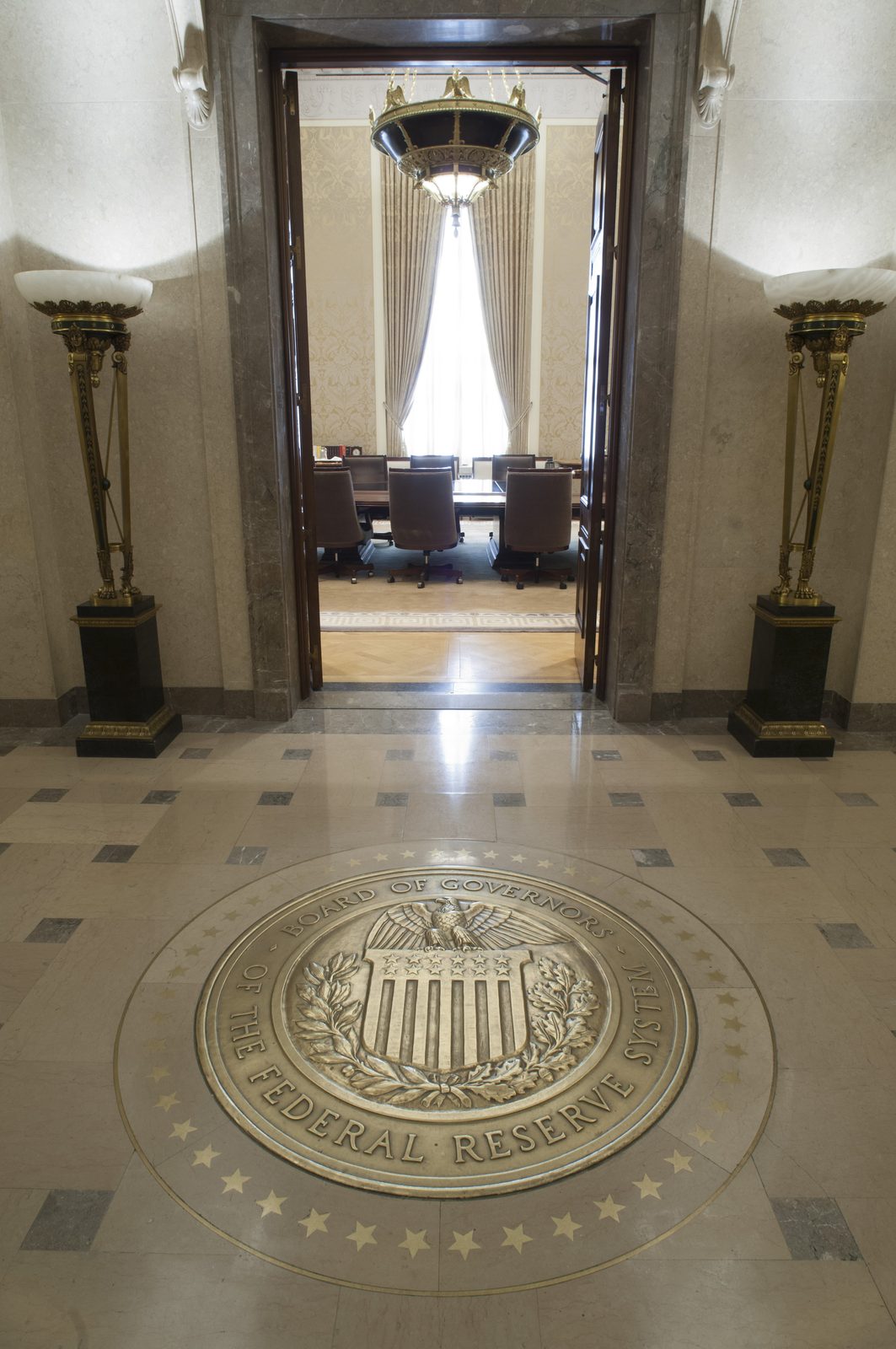 Looking through an open marble doorway into the Federal Reserve's Board Room, empty leather chairs around a long table beyond it, with the bronze Seal of the Board of Governors of the Federal Reserve System set into the floor in the foreground