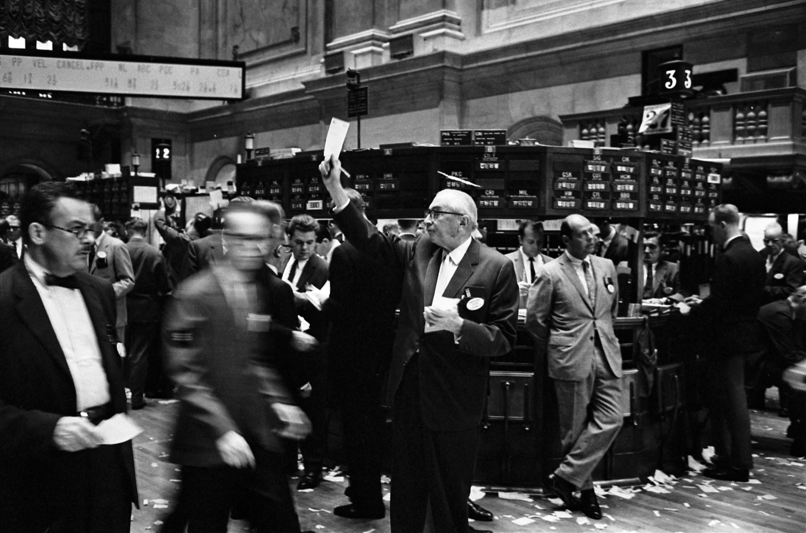Black and white photograph of stock brokers working the trading floor at the New York Stock Exchange in 1963, men in suits crowded around trading posts amid scattered paper