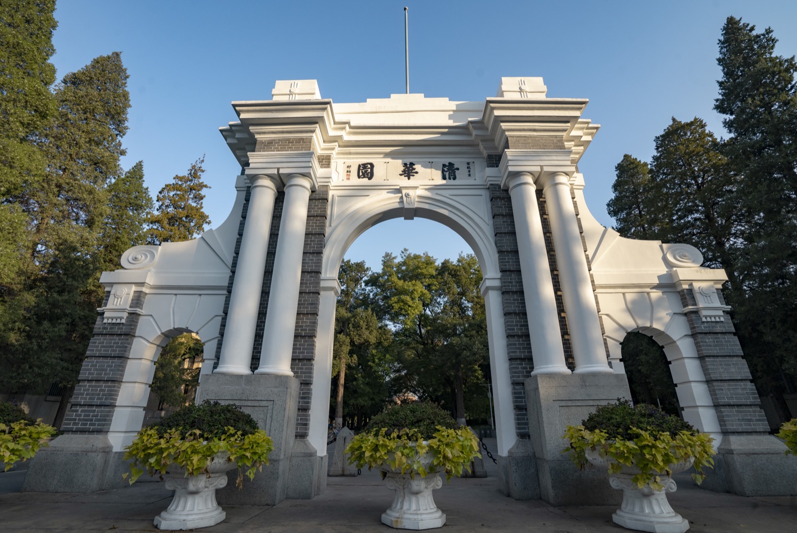 The white stone Second Gate of Tsinghua University in Beijing, a triple-arched Western-classical gateway with paired columns and grey brick, the three characters Qing Hua Yuan carved in red on the central panel, framed by tall green trees under a clear blue sky with three urns of ivy in the foreground