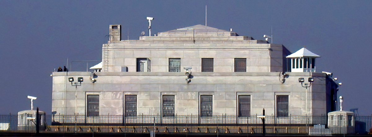 The United States Bullion Depository at Fort Knox, Kentucky — a squat granite vault behind a chain-link perimeter fence, guard towers at its corners, floodlit windows barred