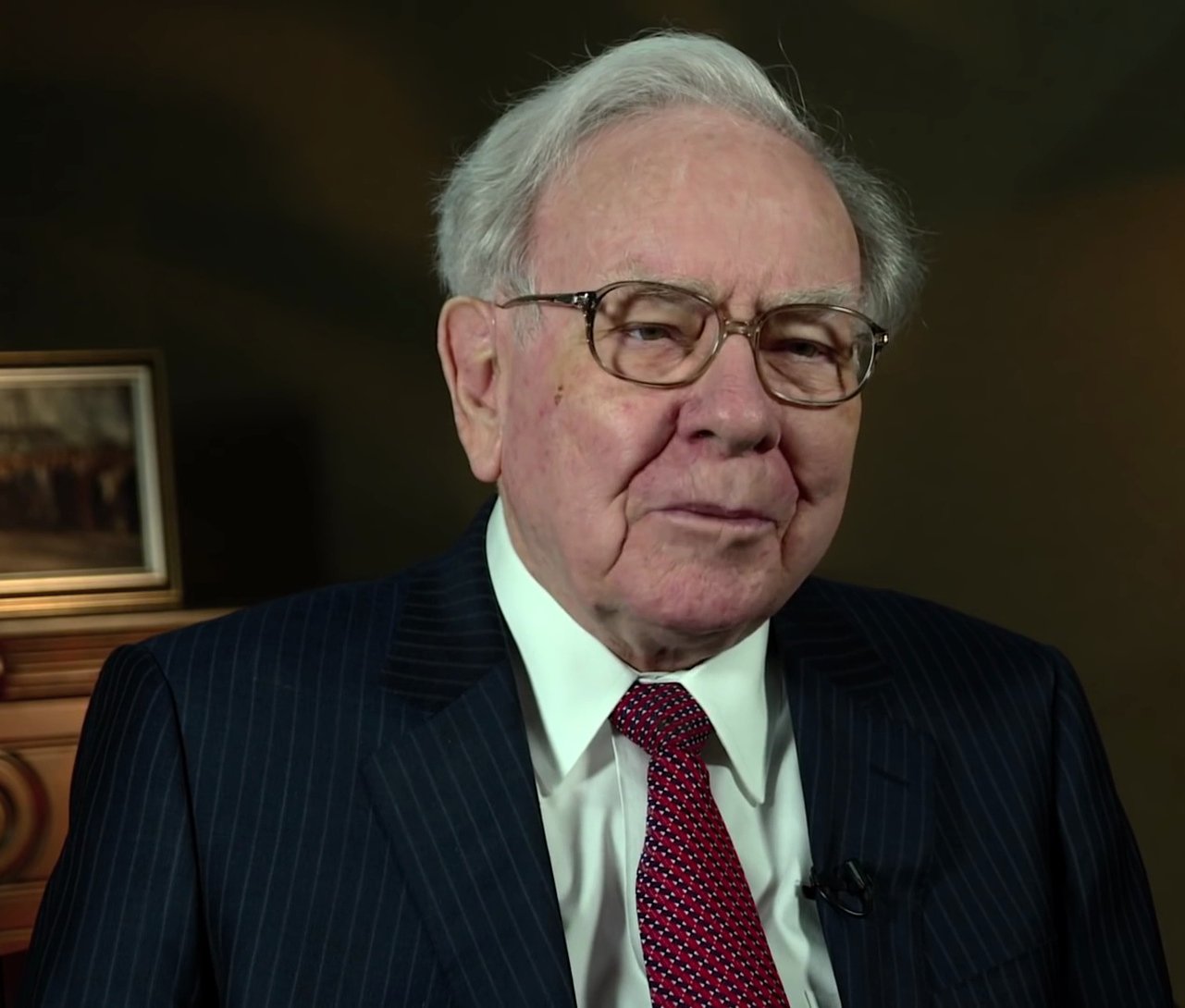 Warren Buffett, an elderly man with white hair and glasses, wearing a dark pinstriped suit and red tie, looking at the camera