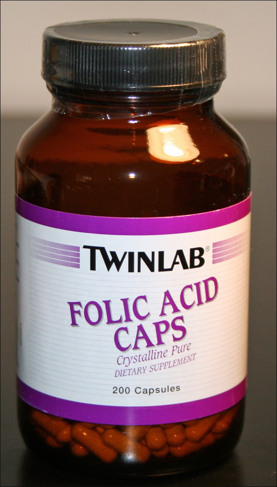 A brown glass supplement bottle labeled "Twinlab Folic Acid Caps, Crystalline Pure, Dietary Supplement, 200 Capsules," with loose orange capsules visible at the base
