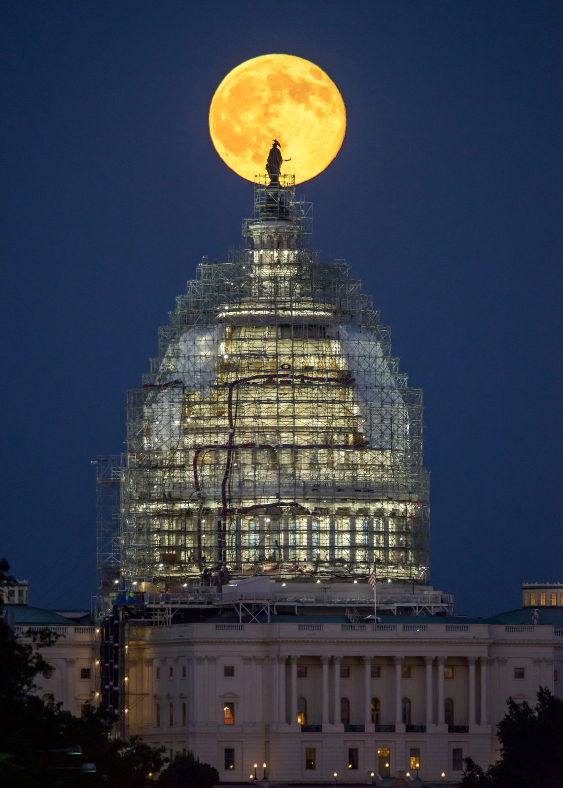 A full moon rising into a dark blue sky behind the illuminated dome of the U.S. Capitol at night