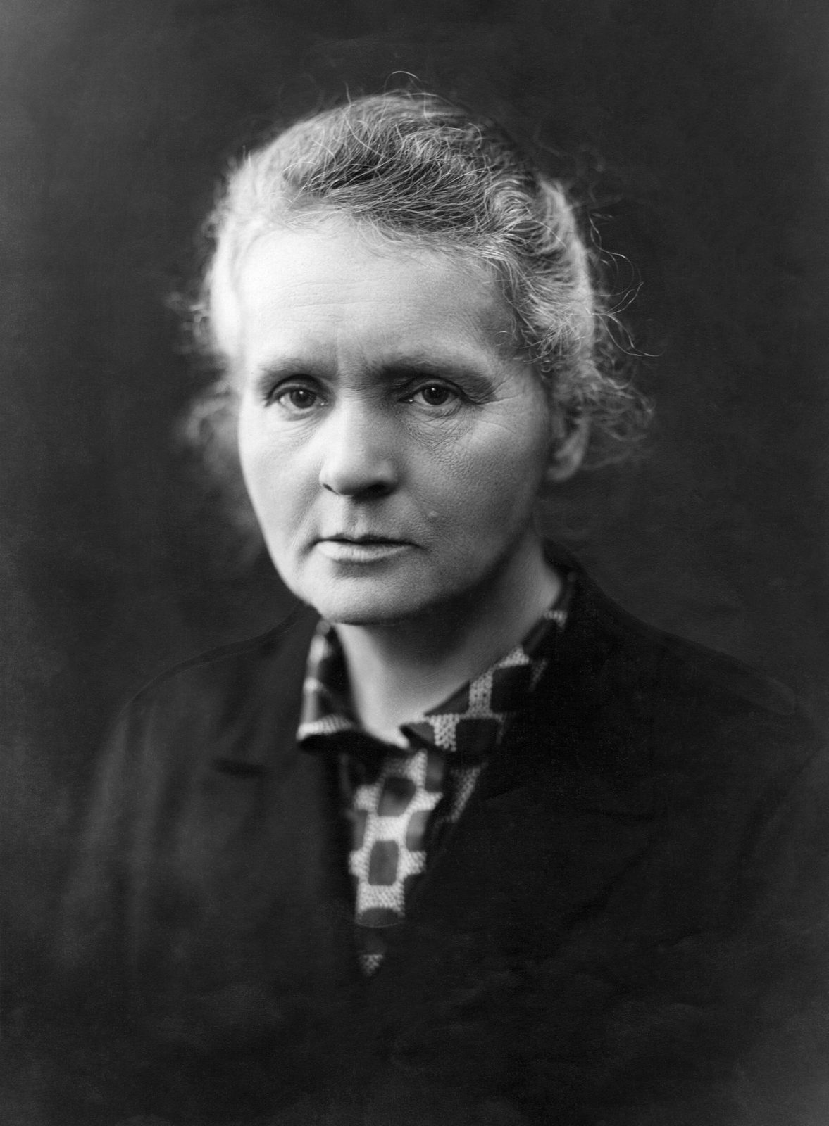 A black-and-white studio portrait of Marie Curie in her fifties, seated, hands folded, in a dark dress, looking slightly off-camera