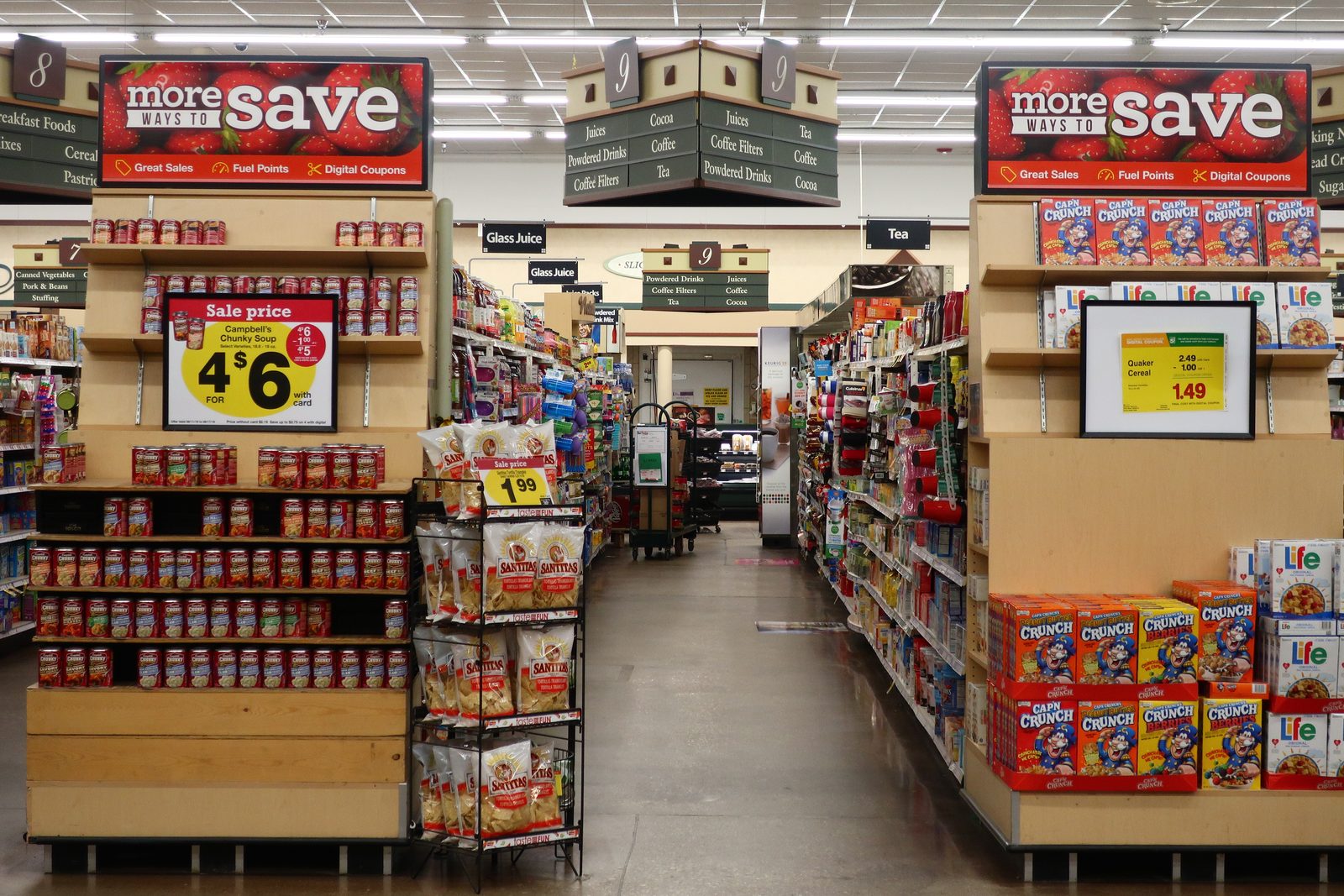 The centre aisle of an American supermarket, looking down a polished floor between two end-cap displays — stacked cans of condensed soup and a rack of bagged tortilla chips on the left, boxed breakfast cereal on the right, with overhead signs reading Juices, Coffee, Tea, Powdered Drinks and Cocoa