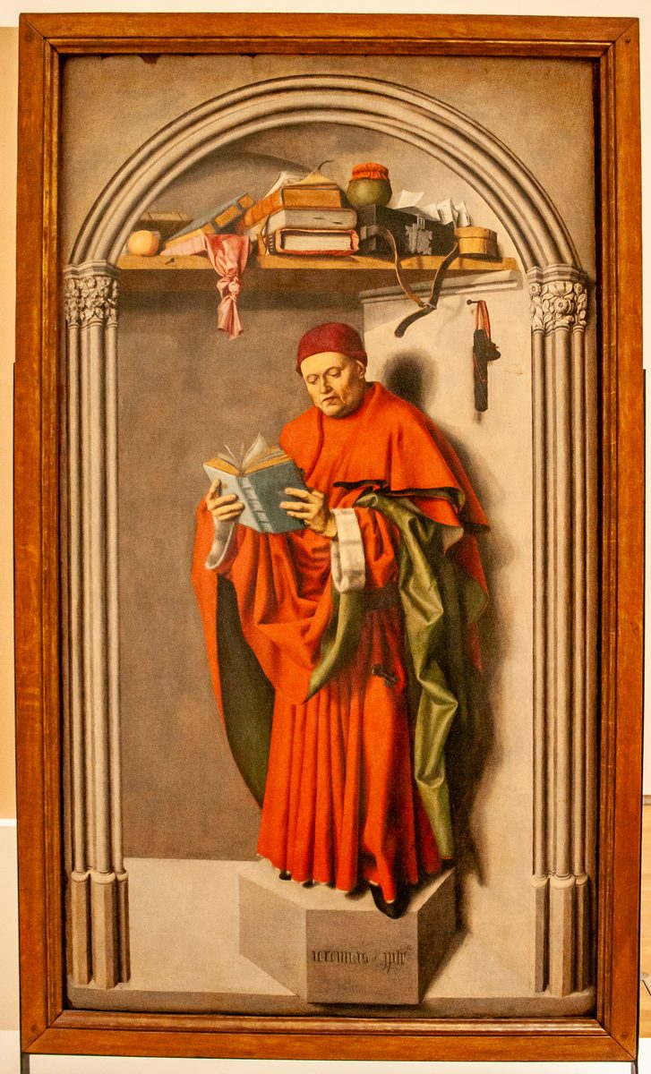 A bearded man in a scarlet cardinal's robe and cap stands reading intently from a small open blue book, framed inside a carved Gothic stone archway on a pedestal; a shelf above him holds books, papers, a quill and a small pot, with a knife and pouch hanging on the wall beside him