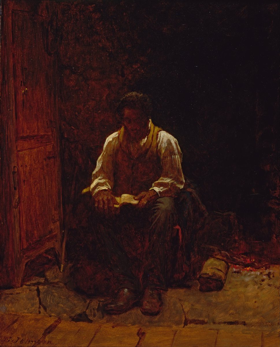 A dark interior. A Black man in a white shirt and dark waistcoat sits alone on a low seat, head bent, holding a small open book in both hands and reading it. A plank door stands open behind him on the left. Embers glow on the floor at the lower right. The only light in the picture falls on his shirt and on the open page.