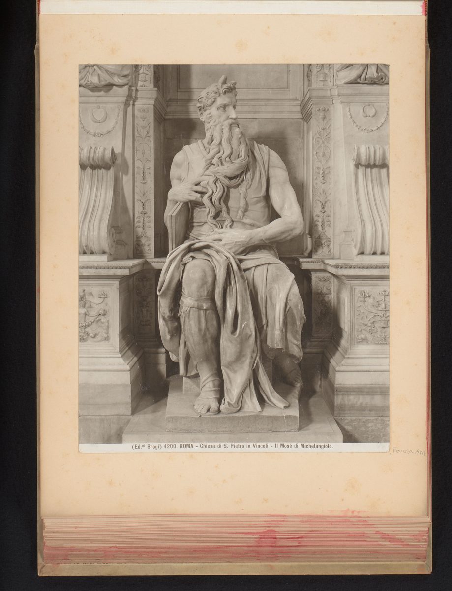A vintage sepia-toned photograph, mounted in an old album with the album's edge visible around it, of a seated marble statue of an elderly bearded man. He wears a loose robe and holds the tablets of the law under one arm while his other hand rests in his long, flowing beard. Two small horn-like protrusions rise from the hair at his temples. He sits in an ornate marble niche flanked by carved pilasters. A caption below the image reads, in Italian, 'Roma — Chiesa di S. Pietro in Vincoli — Il Mosè di Michelangiolo.'