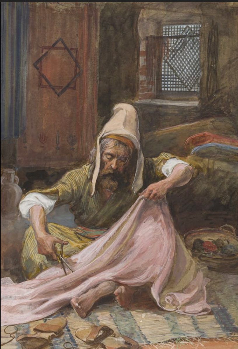 A gouache painting of an elderly bearded man in a pointed hood and striped robe, seated cross-legged on a woven mat, cutting a large piece of pale rose-colored cloth with scissors. He holds the fabric taut with one hand while cutting with the other; sandals and loose threads lie on the mat before him. Behind him, a woven hanging with a star-shaped pattern covers part of a stone wall, a latticed window lets in light, and a basket of colored yarn or dye sits at his side.