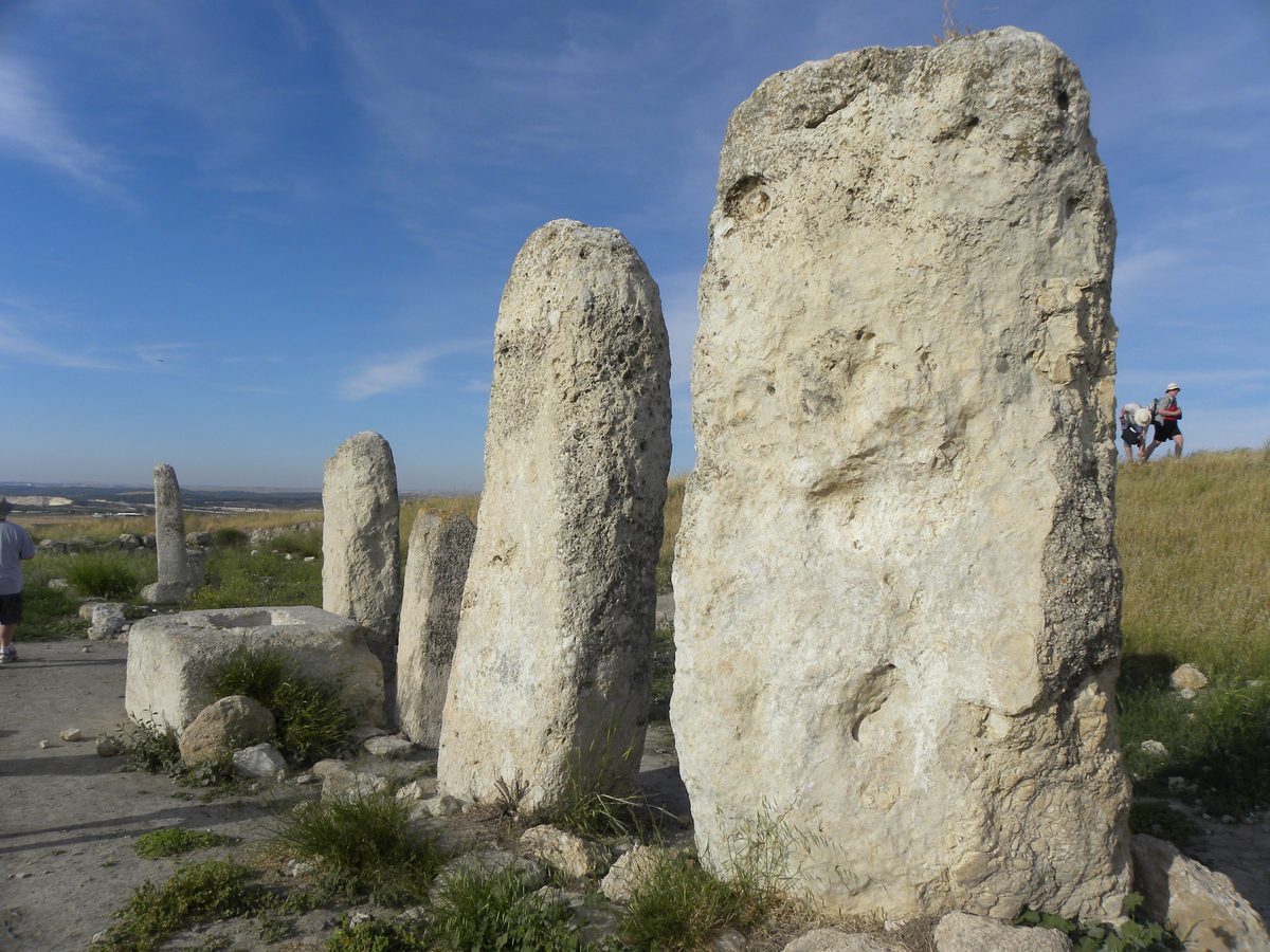 A photograph of a row of large upright limestone monoliths standing in dry grass under a pale sky, the tallest roughly three metres high, spaced several paces apart along a low ridge. Their surfaces are rough and unworked; none is carved or shaped into a figure.