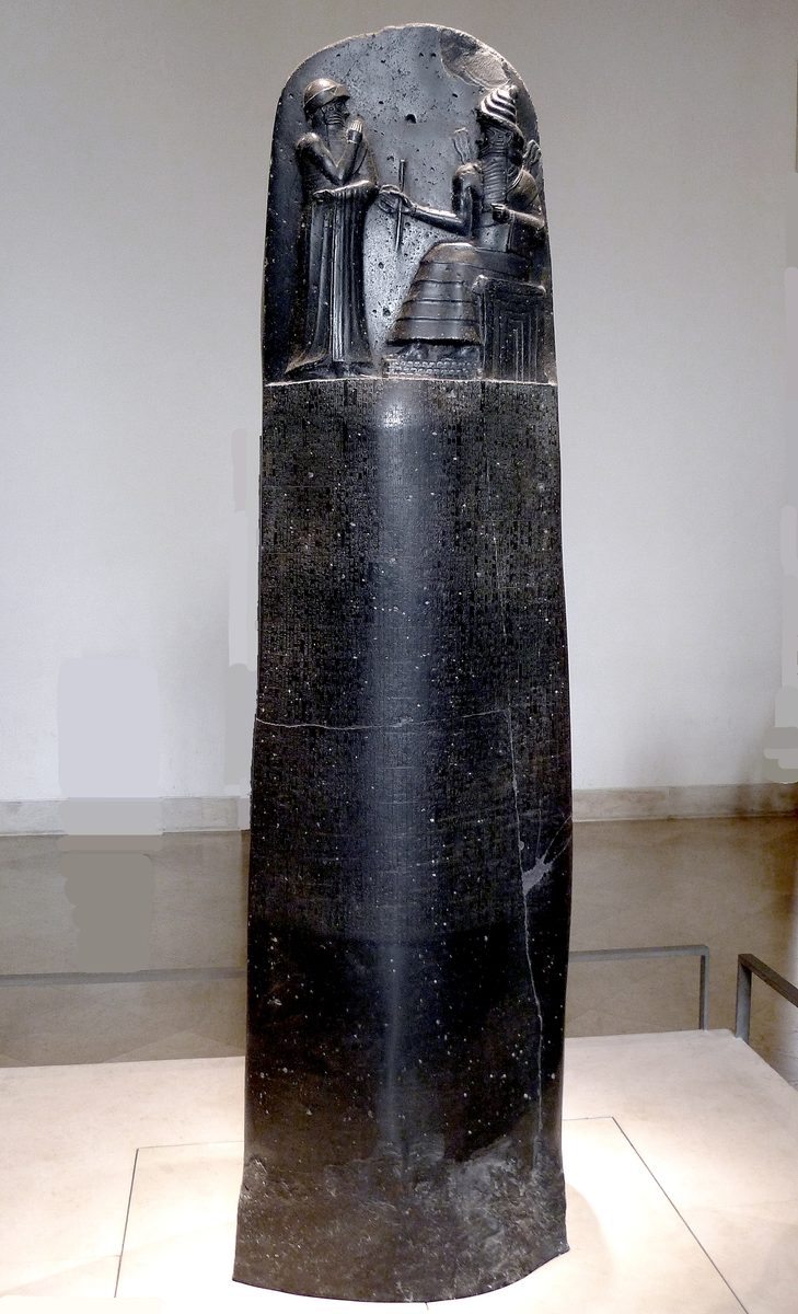 The upper portion of a tall, rounded black basalt stele. A relief carving shows a seated, bearded god on a throne, flames rising from his shoulders, extending a rod and ring toward a standing bearded king who faces him with one arm raised. Below the relief, dense columns of cuneiform script cover the remainder of the stone.