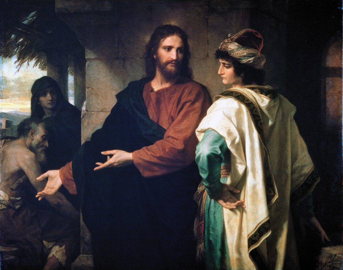 Christ stands at the centre in a red robe and dark blue mantle, turning toward a young man on the right who is dressed in a heavy cream-and-gold cloak over a green sleeve, with a jewelled turban; the young man looks down, hands clasped in front of him. Christ's right hand is open and extended away from him, toward the lower left, where a thin, bare-shouldered old beggar sits on the ground with his palm held out, a woman in a dark hood standing behind him. Stone walls and an archway frame them, with a pale sky visible at the far left
