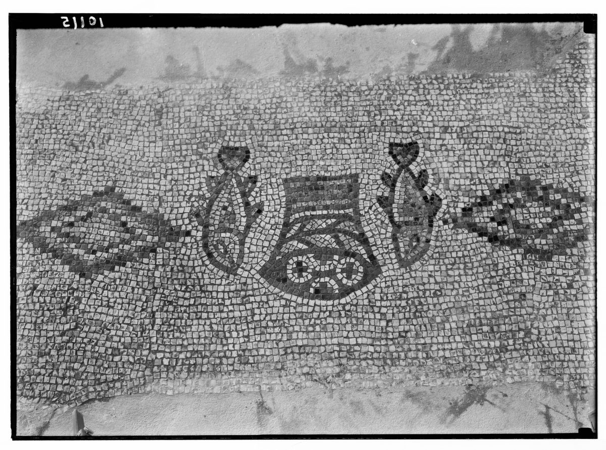 A black-and-white archival photograph of an ancient floor mosaic made of small square stone tesserae. At the center is a woven basket viewed from above, filled with round loaves of bread arranged in a cross pattern. A single fish, rendered in dark stone against the lighter background, flanks the basket on each side, both facing inward toward it. The surrounding mosaic field is a plain repeating pattern of small light-colored tiles, with scattered dark stains and shadows across the surface from age and weathering.