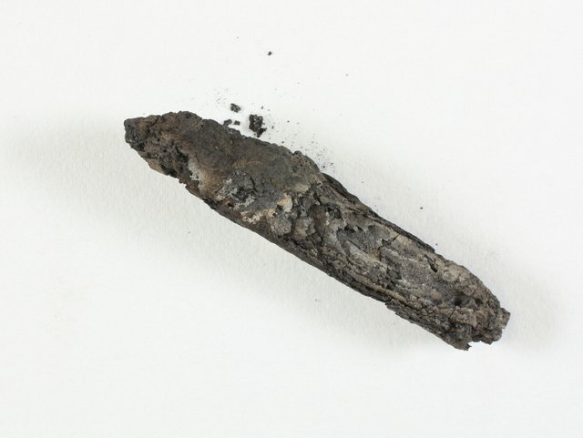 A small, blackened, cylinder-shaped lump of charred, crumbling parchment, tightly rolled and badly burned, photographed against a plain white background.