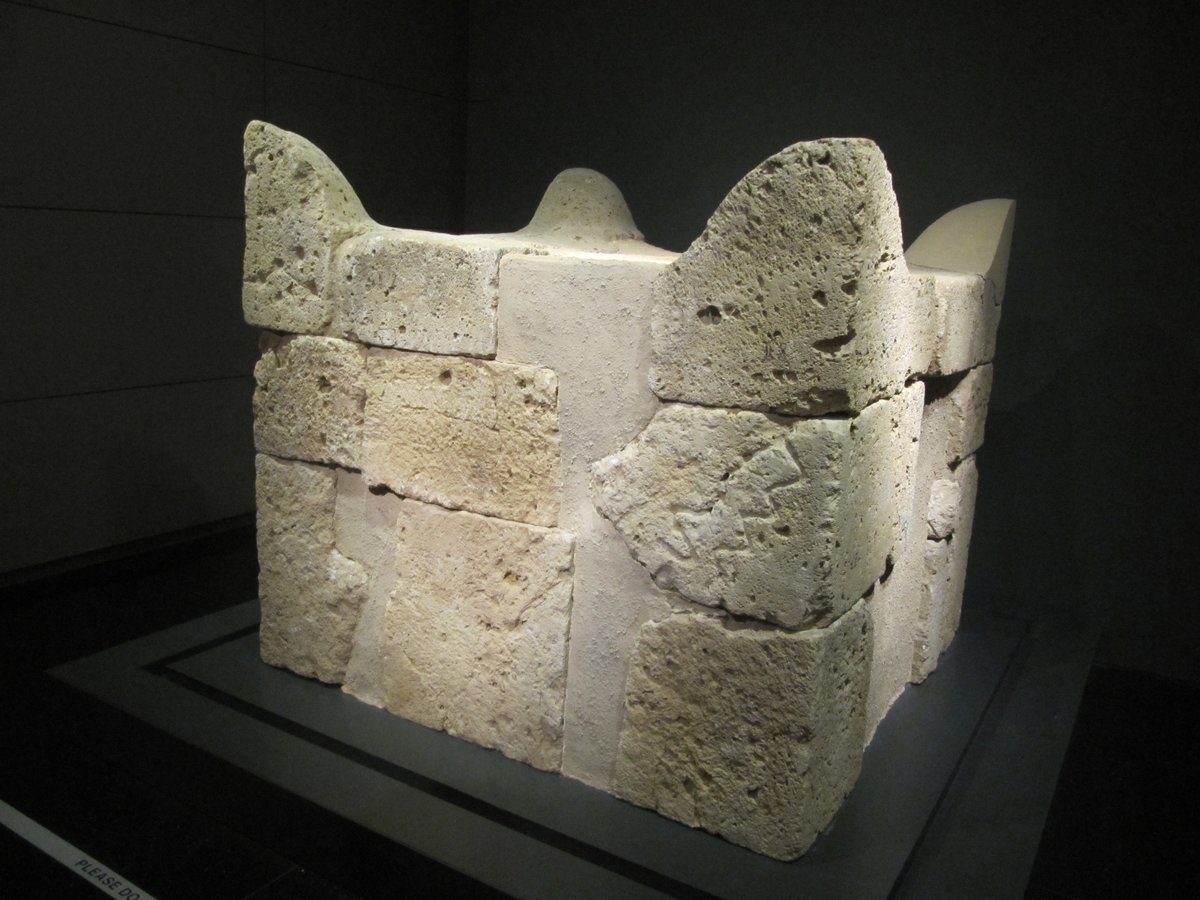 A reassembled ancient altar built of large rectangular sandstone blocks, its four upper corners rising into distinct curved horn-shapes, displayed on a low platform in a dimly lit museum gallery.