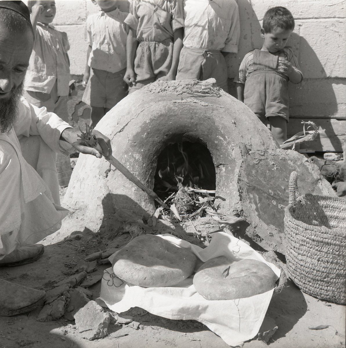 A black-and-white photograph of an elderly bearded man in traditional dress, kneeling beside a dome-shaped clay oven built over an open fire, tending it with a stick. Two round loaves of fresh flatbread rest on a cloth in front of the oven; a group of children stand watching behind him.