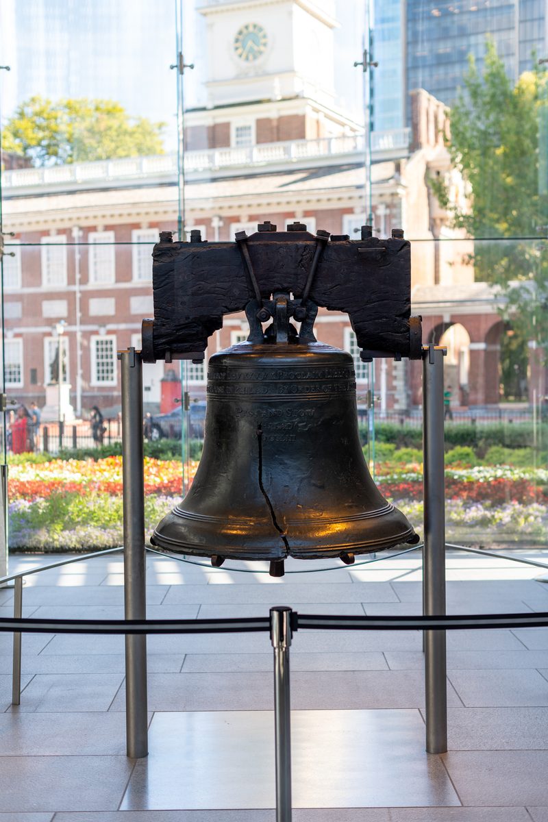 The cracked Liberty Bell hangs in its glass pavilion, its famous fracture running up through the visible cast inscription. Independence Hall's clock tower is framed behind it through the glass, with a garden of red and yellow flowers in front.