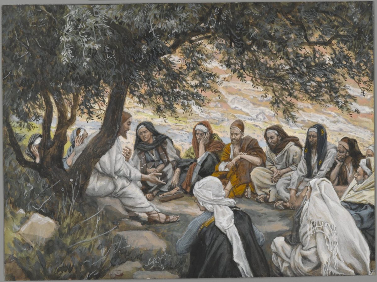 Under the low branches of an olive tree on a bare hillside, Jesus sits at left in plain white and sandals, leaning forward with both hands open as he speaks; the Twelve are arranged in a loose semicircle on the rocky ground around him, wrapped in the everyday robes and head-cloths of the region, several with a hand raised to the face in the posture of men listening hard, a hazy line of hills fading into a pale sky behind them.