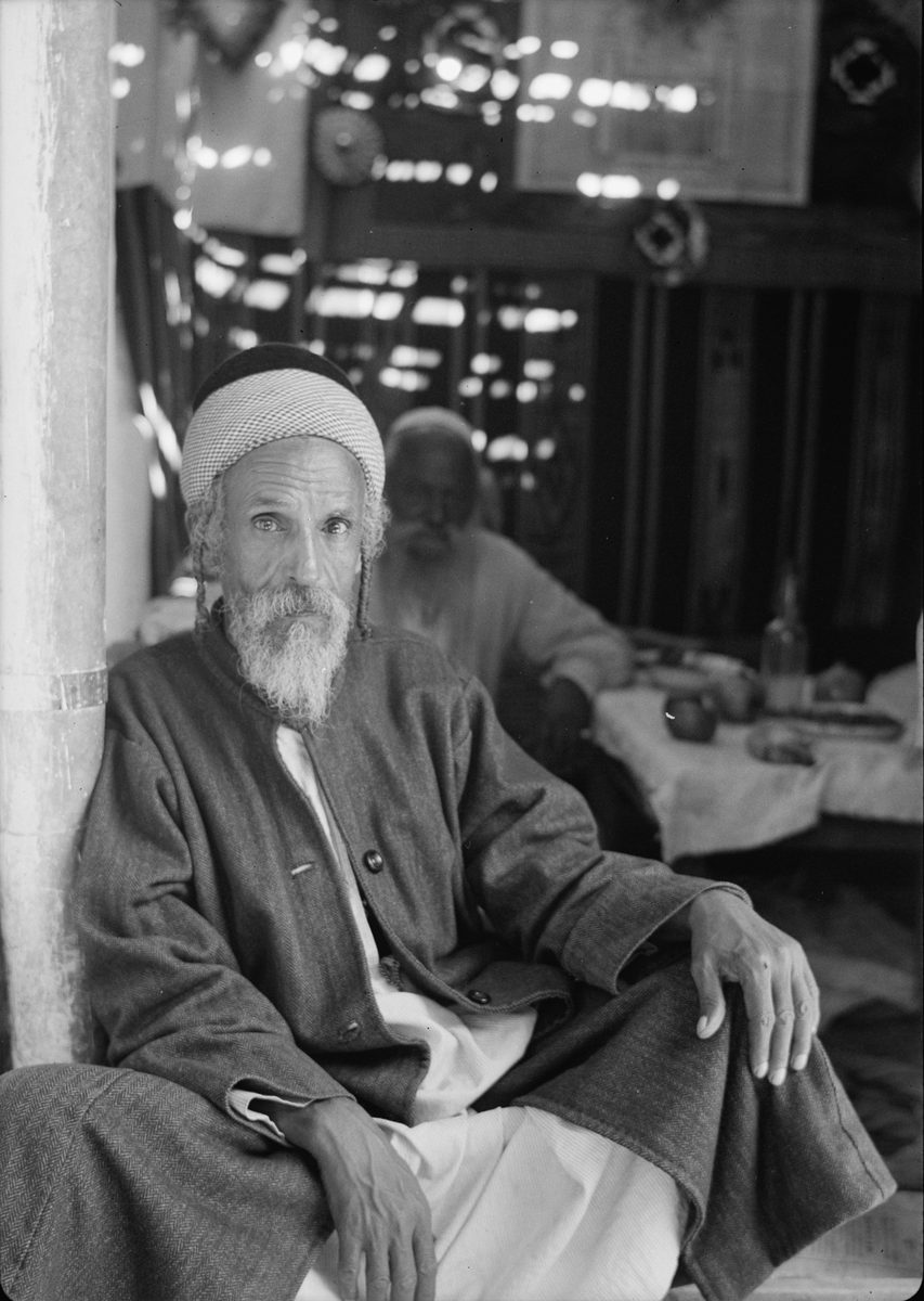 A black-and-white photograph of an elderly bearded man in a striped headwrap and wool jacket, seated in the doorway of a temporary booth. Above him the roof is loosely woven branches, letting dappled light filter through in bright patches across the dim interior. A second man sits further back at a cloth-covered table set with dishes and a bottle. A wooden support post stands at the left edge of the frame.