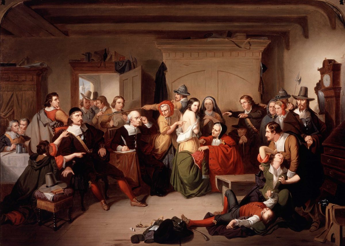 A crowded 17th-century colonial room. At the center, a young woman stands with one shoulder bared while an older woman in a red cloak inspects her skin; magistrates in wide-brimmed hats sit at a table on the left, one writing notes; men in steel helmets and Puritan dress point and gesture around her. In the foreground, a woman appears to have collapsed into a fit and is held up by another figure. A tall grandfather clock stands at the right.
