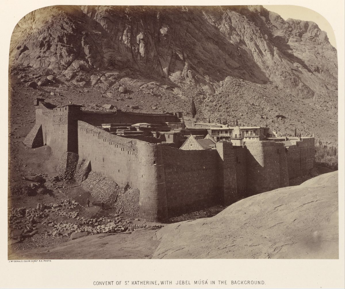 A sepia-toned 19th-century photograph of a massive fortified stone monastery compound at the base of a steep, rocky mountain that fills most of the background. Thick crenellated walls enclose a cluster of stone and timber buildings; a few tiny human figures stand near the base of the wall, dwarfed by both the fortification and the mountain behind it. A printed caption below the image reads: CONVENT OF ST KATHERINE, WITH JEBEL MUSA IN THE BACKGROUND.