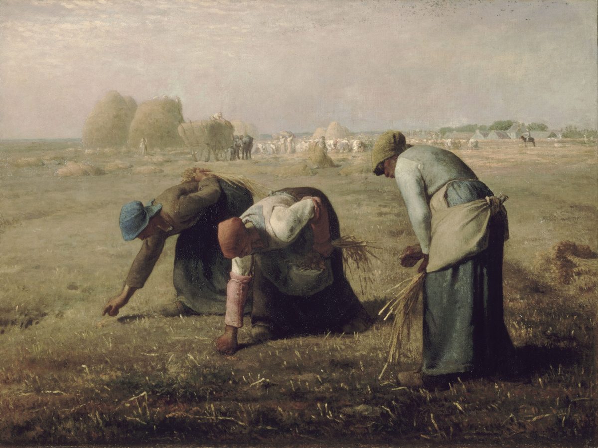 Three peasant women, bent low, gather loose stalks of wheat from a stubbled field in the foreground. The nearest woman, in a green cap, stands more upright with a sheaf tucked under her arm; the other two are bent nearly double, faces hidden. In the hazy distance behind them, a much busier harvest scene unfolds under a pale sky: haystacks, a loaded wagon, a crowd of workers and a mounted overseer.
