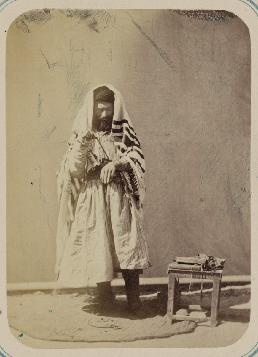 A sepia photograph of a bearded man standing against a bare plastered wall. A striped prayer shawl is draped over his head and shoulders; long twisted fringes hang in heavy bundles at each hip. His left arm is wrapped in the leather straps of a phylactery and his head is bowed. He stands on a small rug beside a low wooden stand holding books.