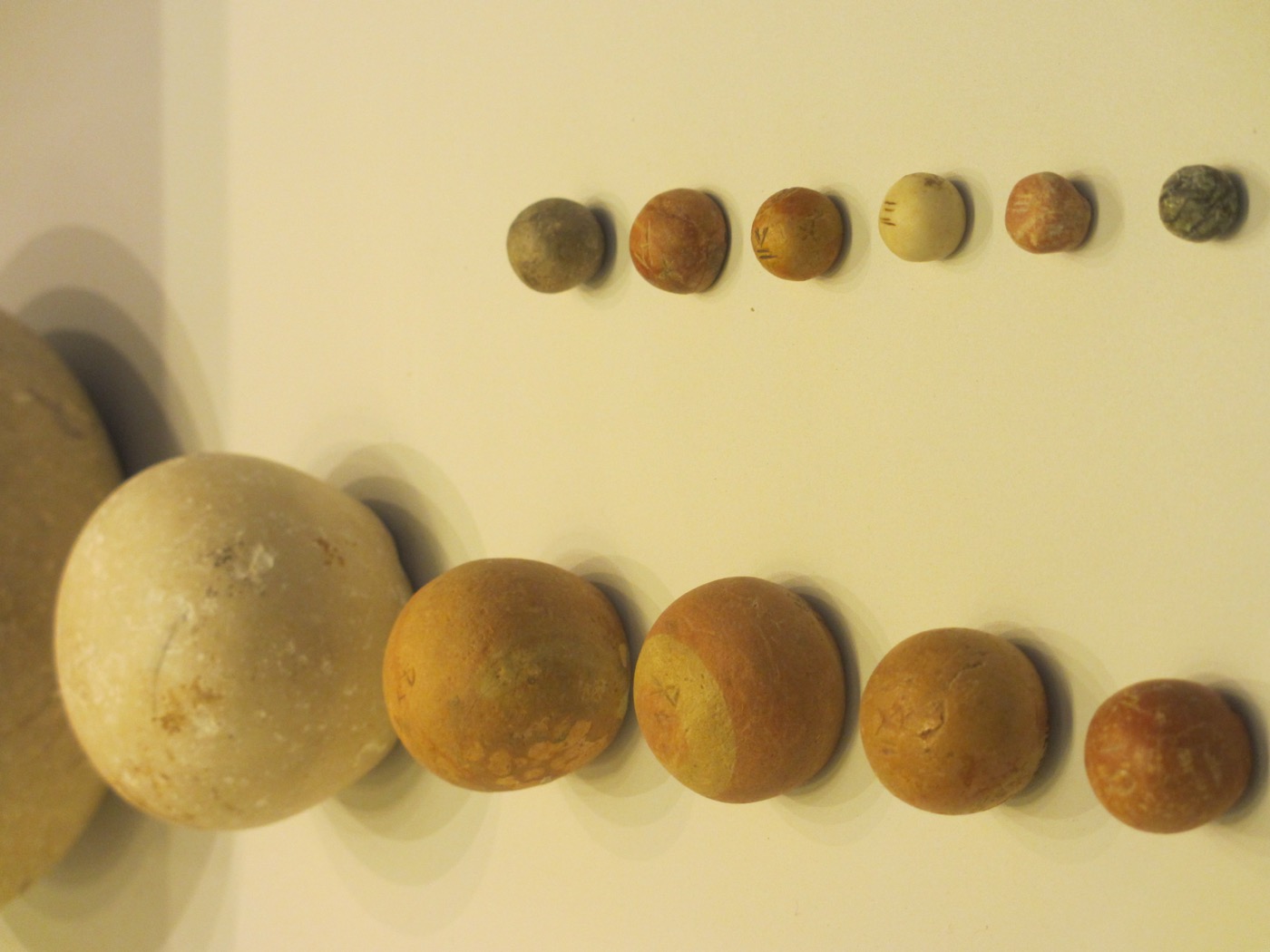 A museum display of polished dome-shaped stone weights on a cream surface, arranged in two columns. At the very top edge a sixth stone is cut off by the frame; below it the left column runs from a large pale limestone dome down through four progressively smaller ochre domes. The right column holds six much smaller weights, from pea-sized down to little more than a bead. Several of the stones carry shallow incised marks scratched into their flat upper faces.