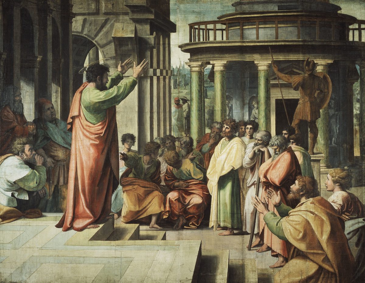 A large tapestry-cartoon painting. A bearded man in a green tunic and orange-red cloak, with a faint halo, stands on a raised stone platform with both arms lifted and hands open, mid-gesture, addressing a crowd below and around him. The listeners are a mixed group in robes of green, orange, and white, some seated on the steps, some standing, with varied expressions — attentive, skeptical, one man crouched with his head in his hands. Behind them rises a grand classical rotunda with green marble columns and a bronze statue of an armored, spear-carrying figure on a pedestal.