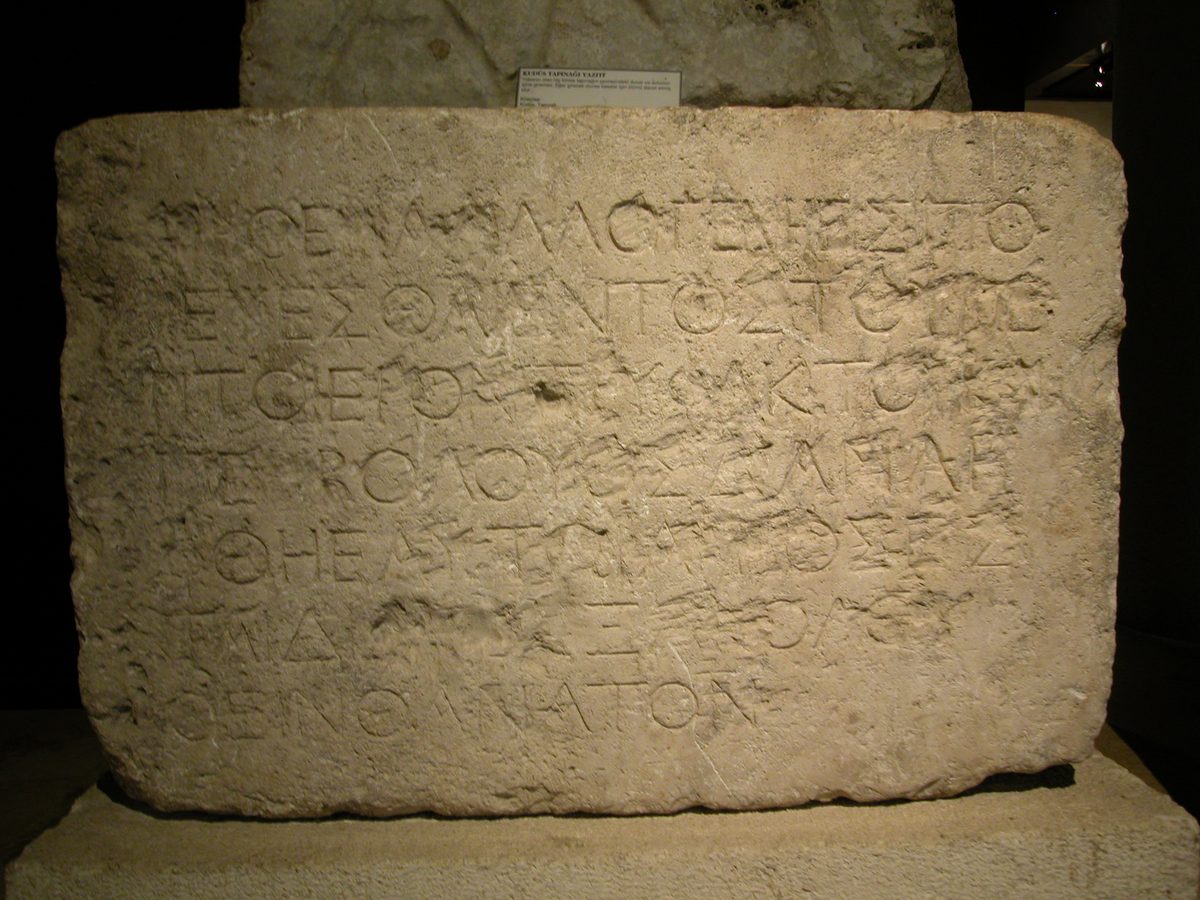 A photograph of a large tan limestone block on a museum pedestal, lit against a dark background. Seven lines of ancient Greek capital letters are carved across its face, worn and pitted with age but still legible in most places. A small museum label is visible on a stand behind the stone.