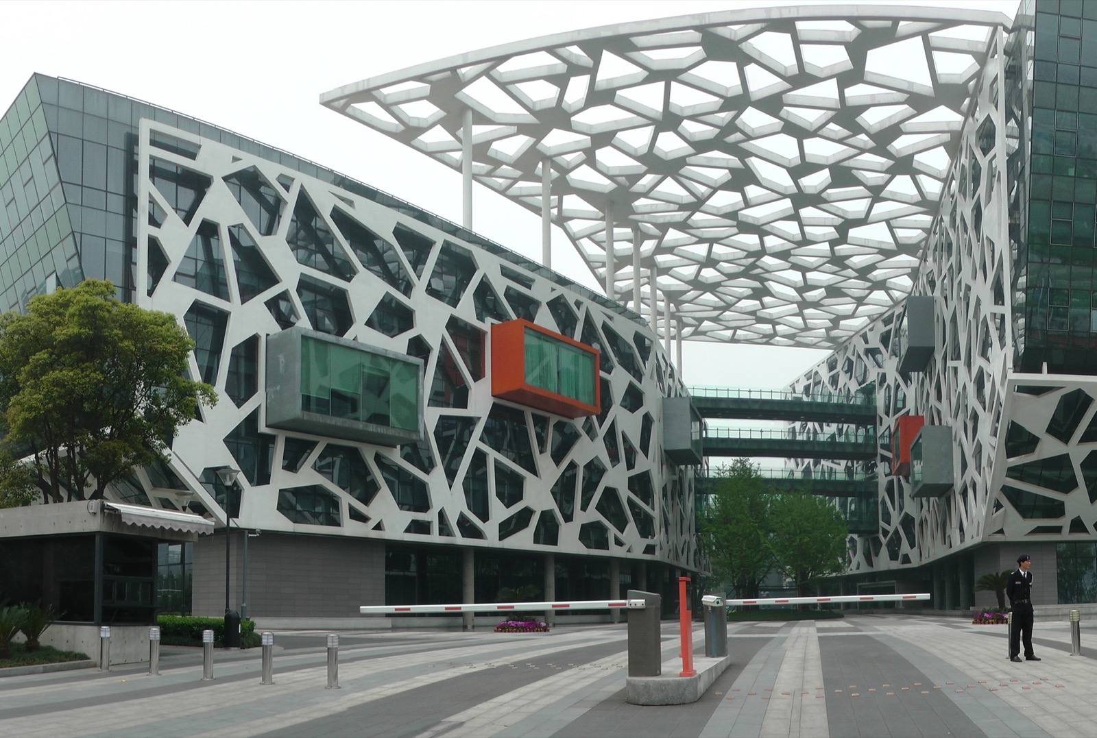 The entrance plaza of Alibaba Group's headquarters campus in Hangzhou, China — low glass office blocks wrapped in a white concrete lattice of irregular polygons, with a matching perforated canopy floating overhead on slender columns, orange and grey box-shaped meeting rooms cantilevered out of the facade, and a security barrier across the empty forecourt