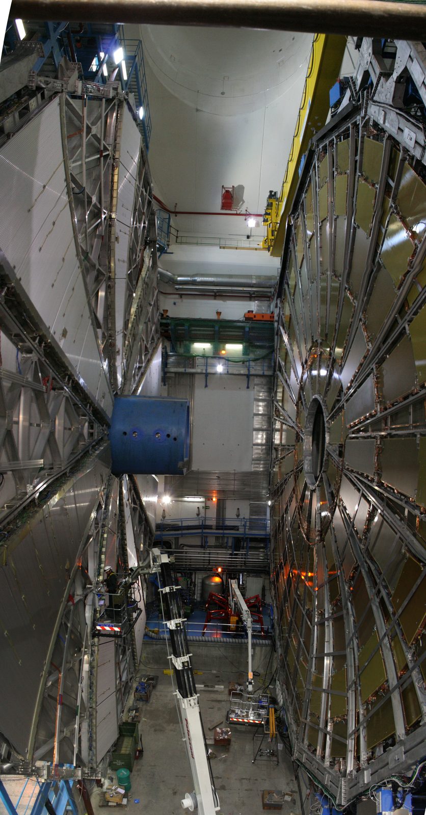 The ATLAS particle detector under construction inside its underground cavern at CERN, seen from above between its two wheel-shaped end-caps, with scaffolding, cranes and a blue crane platform visible around the silver and gold detector components