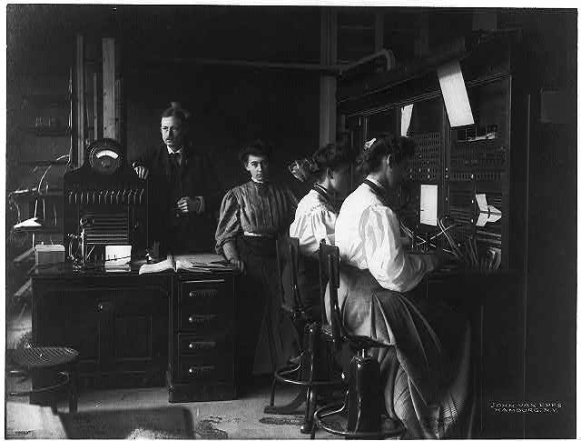 A telephone exchange office around 1900, two women operating switchboards while two men attend nearby equipment, one leaning in close to check a line