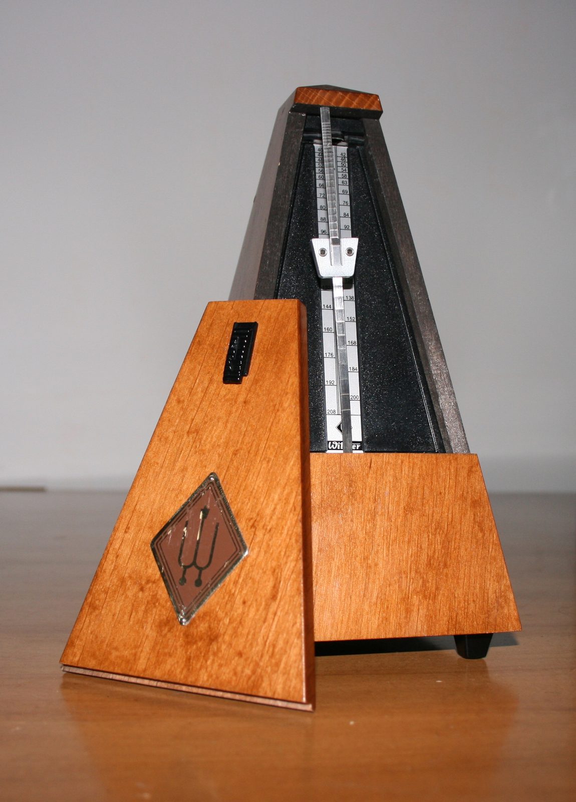 A close-up photograph of a traditional wooden pyramid metronome, its tall pendulum arm tipped with a sliding brass weight, set against a plain background
