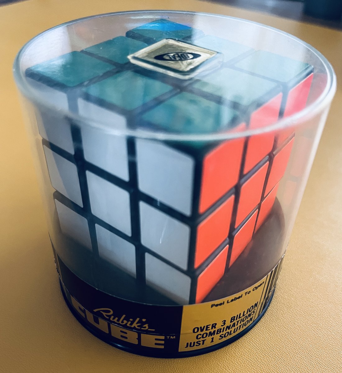 An original 1980 Rubik's Cube inside Ideal Toy's clear cylindrical plastic canister, sitting on a tan table. The white, red and blue faces show, and the top center square carries a small gold Ideal sticker. The black base is labelled Rubik's Cube, with a gold panel reading Over 3 billion combinations, just 1 solution, and a tab reading Peel label to open.