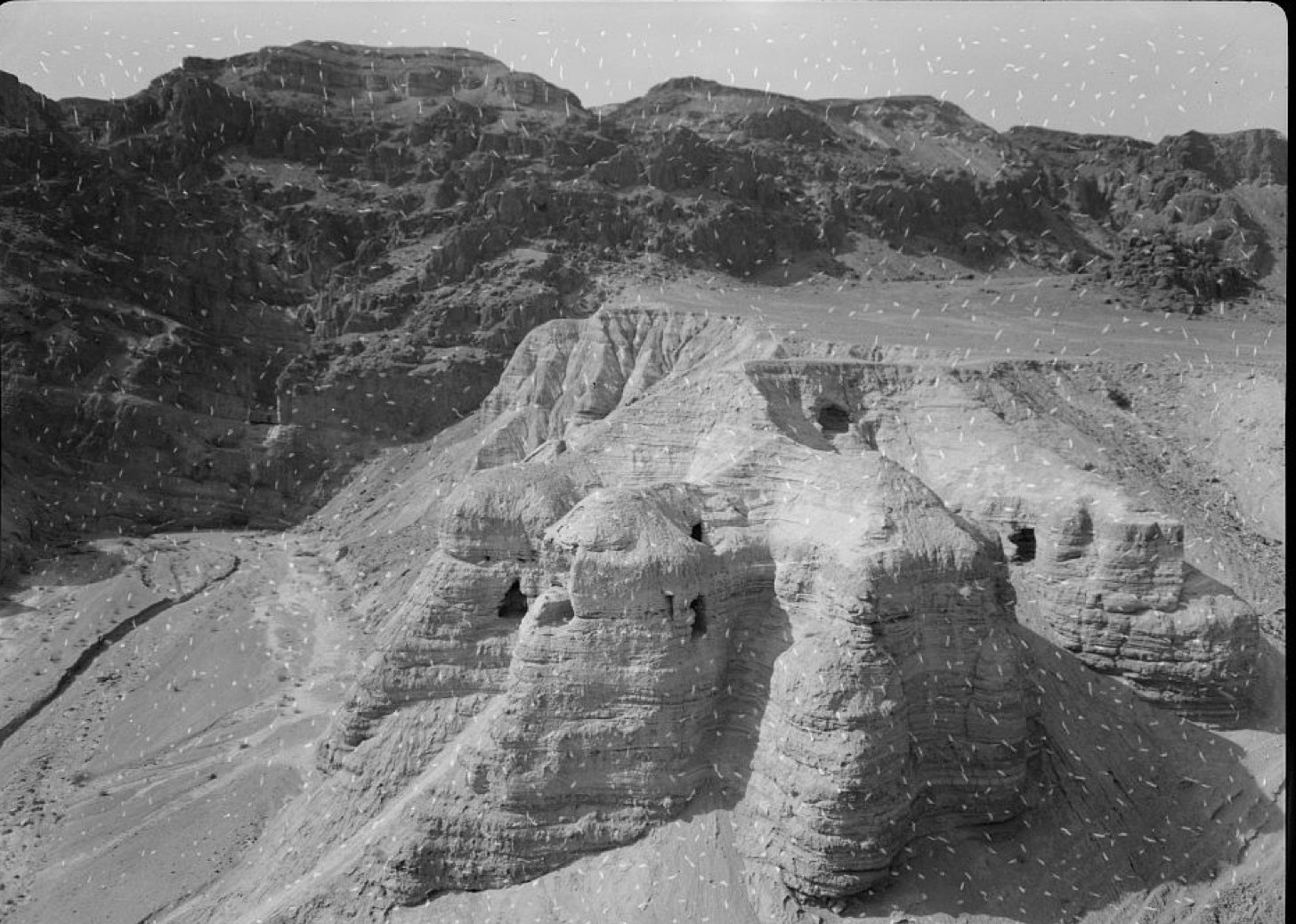 Black-and-white archival photograph of the marl terrace at Qumran in the Judaean desert, with two dark rectangular cave openings cut into a pale eroded cliff of soft sediment in the foreground and the darker rocky escarpment of the Judaean hills rising behind; white speckles from the original negative are scattered across the frame
