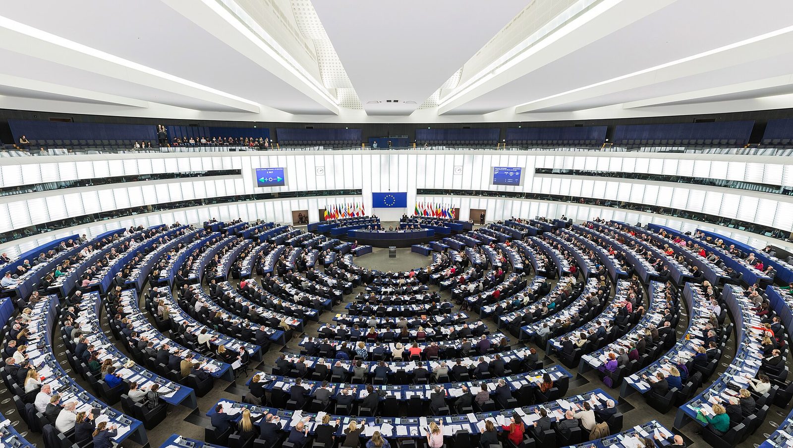 The hemicycle of the European Parliament in Strasbourg seen from the back of the chamber, a wide curved room of blue seats sloping down to the president's rostrum, mostly full of members during a plenary session