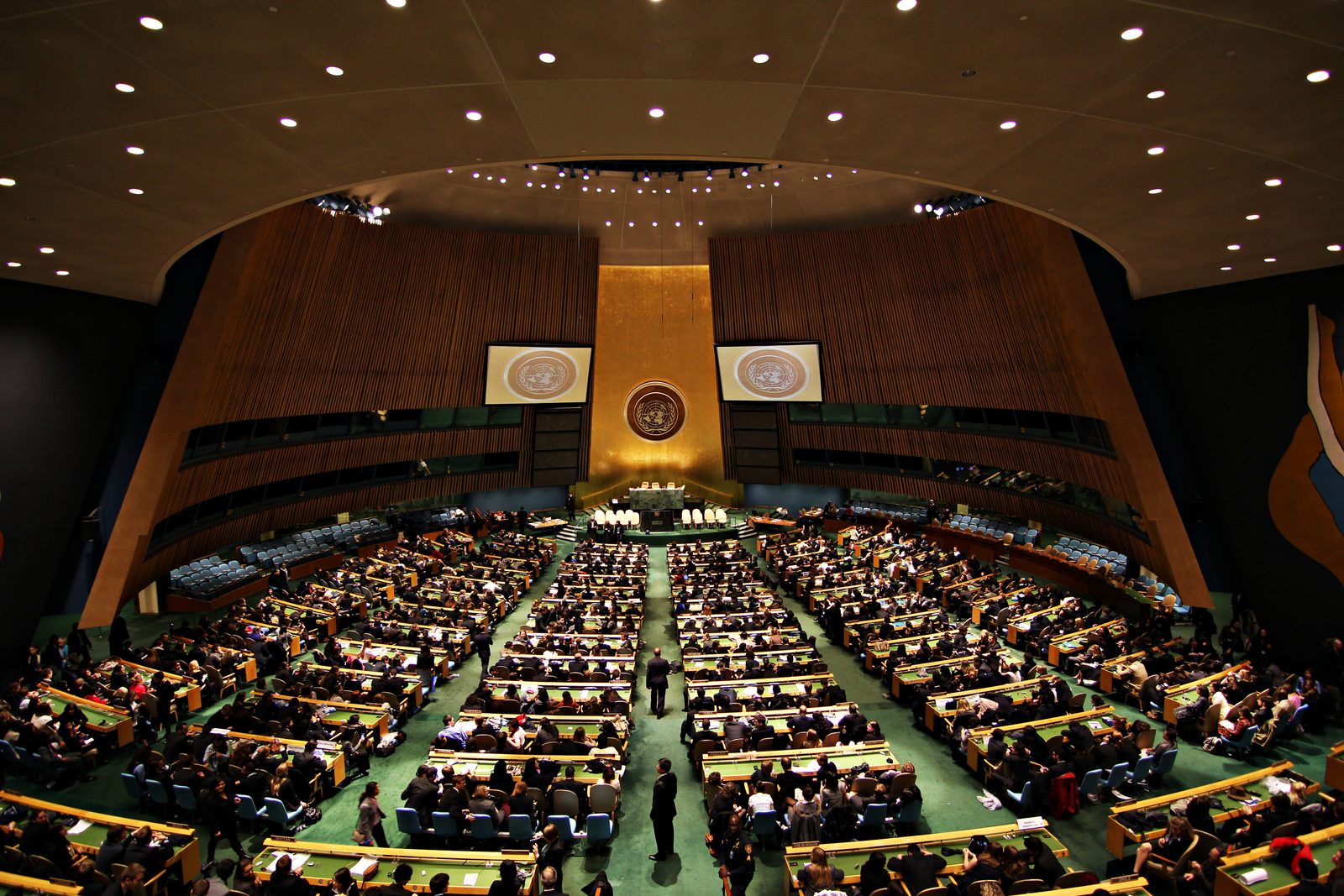 A wide fisheye view inside the United Nations General Assembly Hall in New York, looking down from the upper gallery at rows of delegates seated at green desks, with the gold UN emblem and rostrum at the front of the chamber