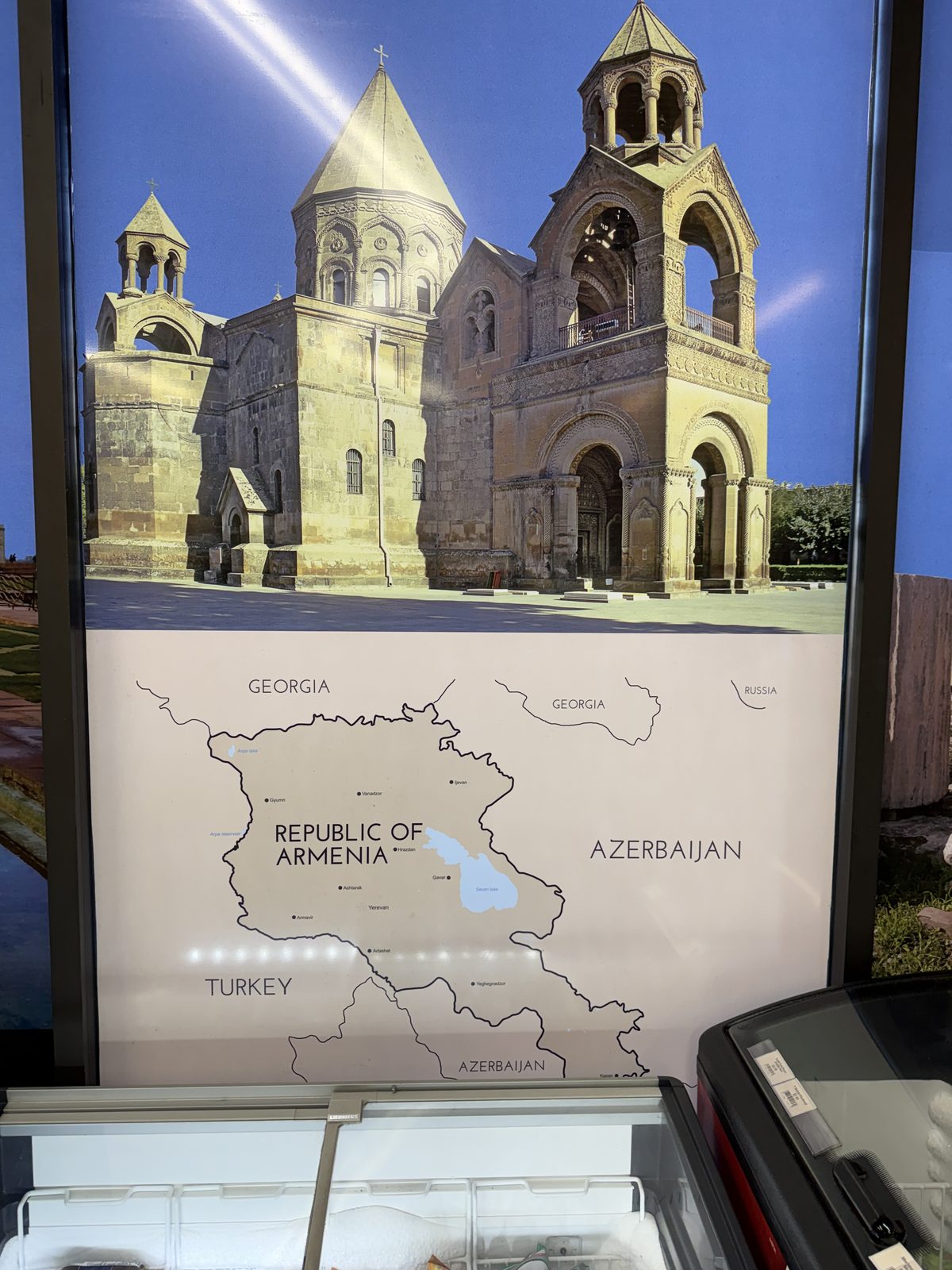 A poster on the wall at Anoush Deli showing the tiered stone bell towers of an Armenian cathedral above a map of the Republic of Armenia bordered by Georgia, Azerbaijan and Turkey, mounted above a chest freezer