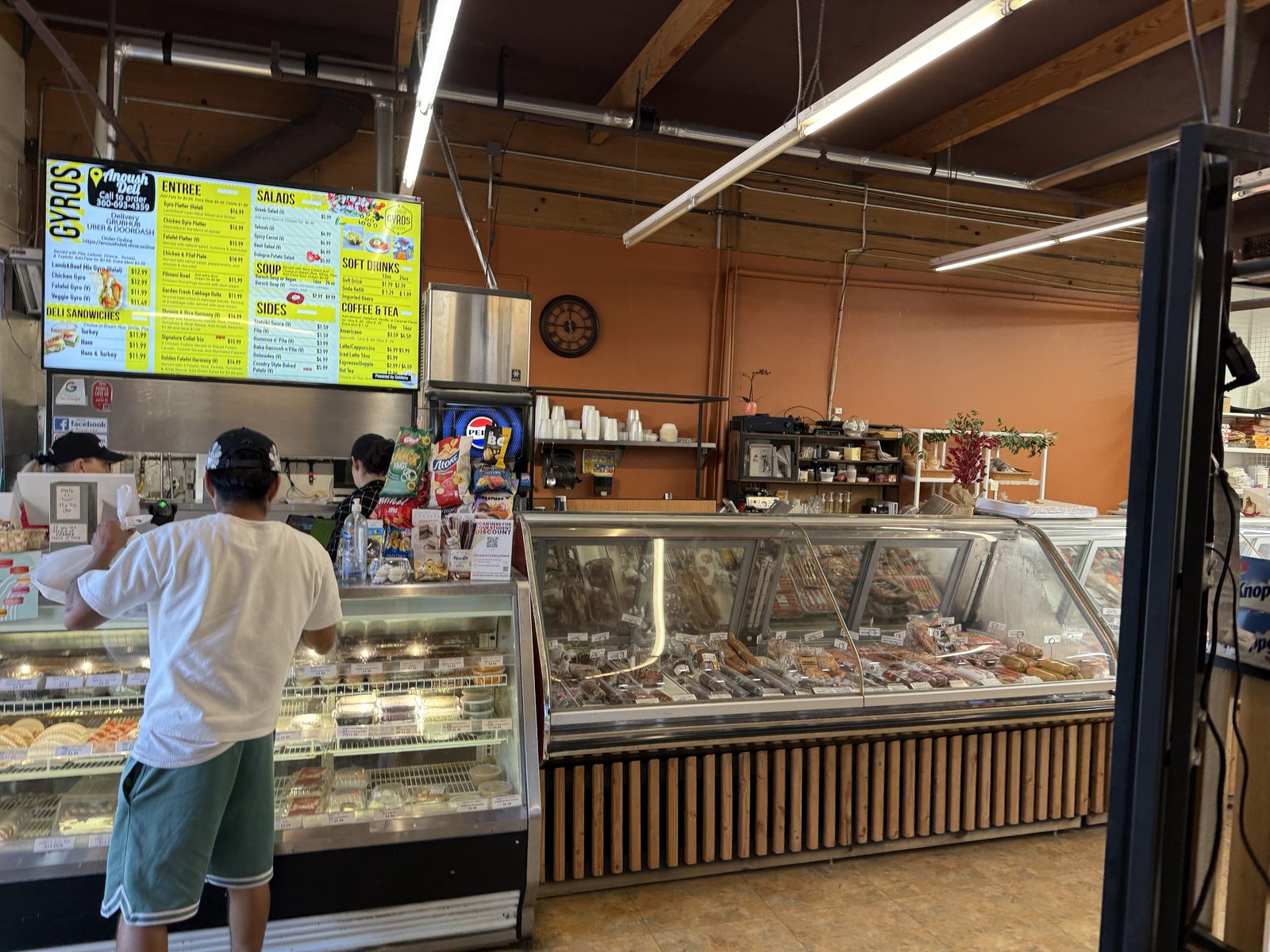 A backlit yellow and light-blue digital menu board reading GYROS above the Anoush Deli logo and phone number, listing a Lamb and Beef Mix Gyro Halal for $12.99 among Entree, Salad, Soup, Sides, Soft Drink and Coffee sections, with a customer in a white t-shirt and cap ordering at the counter below and a long glass deli case of sausages and cured meats to the right