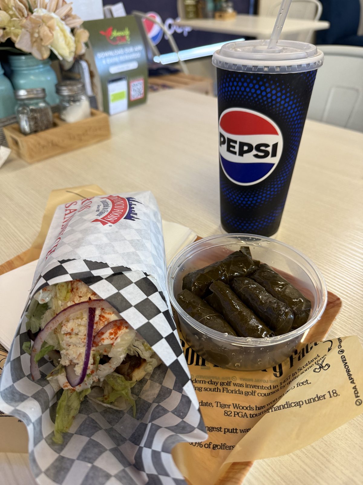 A beef and lamb gyro wrapped in checkered paper, packed with meat, feta, shredded lettuce and red onion under a dusting of red spice, next to a clear container of five stuffed grape leaves and a Pepsi fountain drink with a straw, on a table at Anoush Deli in Vancouver, Washington