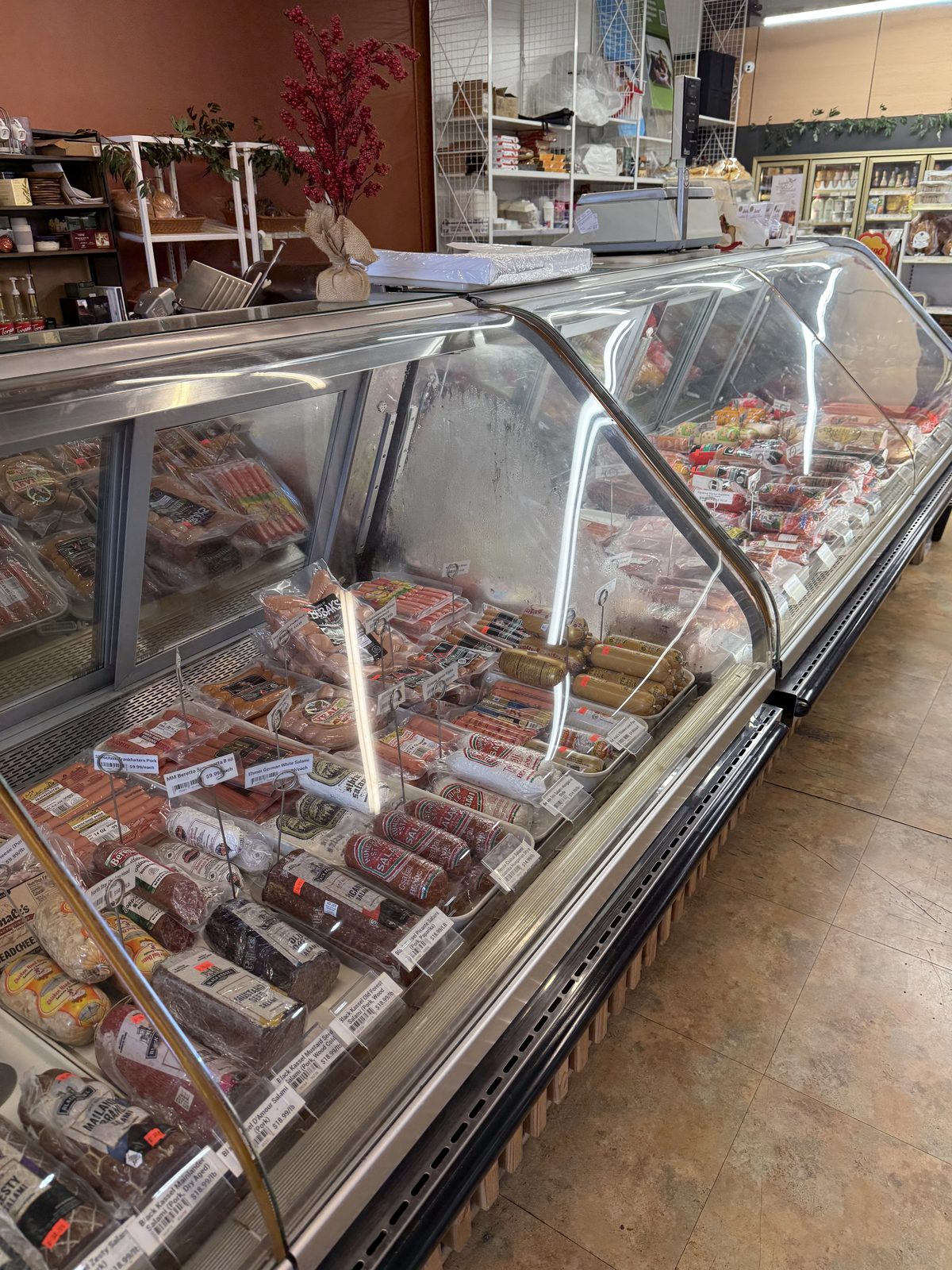 A long curved-glass deli case at Anoush Deli filled with rows of packaged sausages, salami, hot dogs and sliced cold cuts, each with a handwritten price tag, under warm overhead lighting
