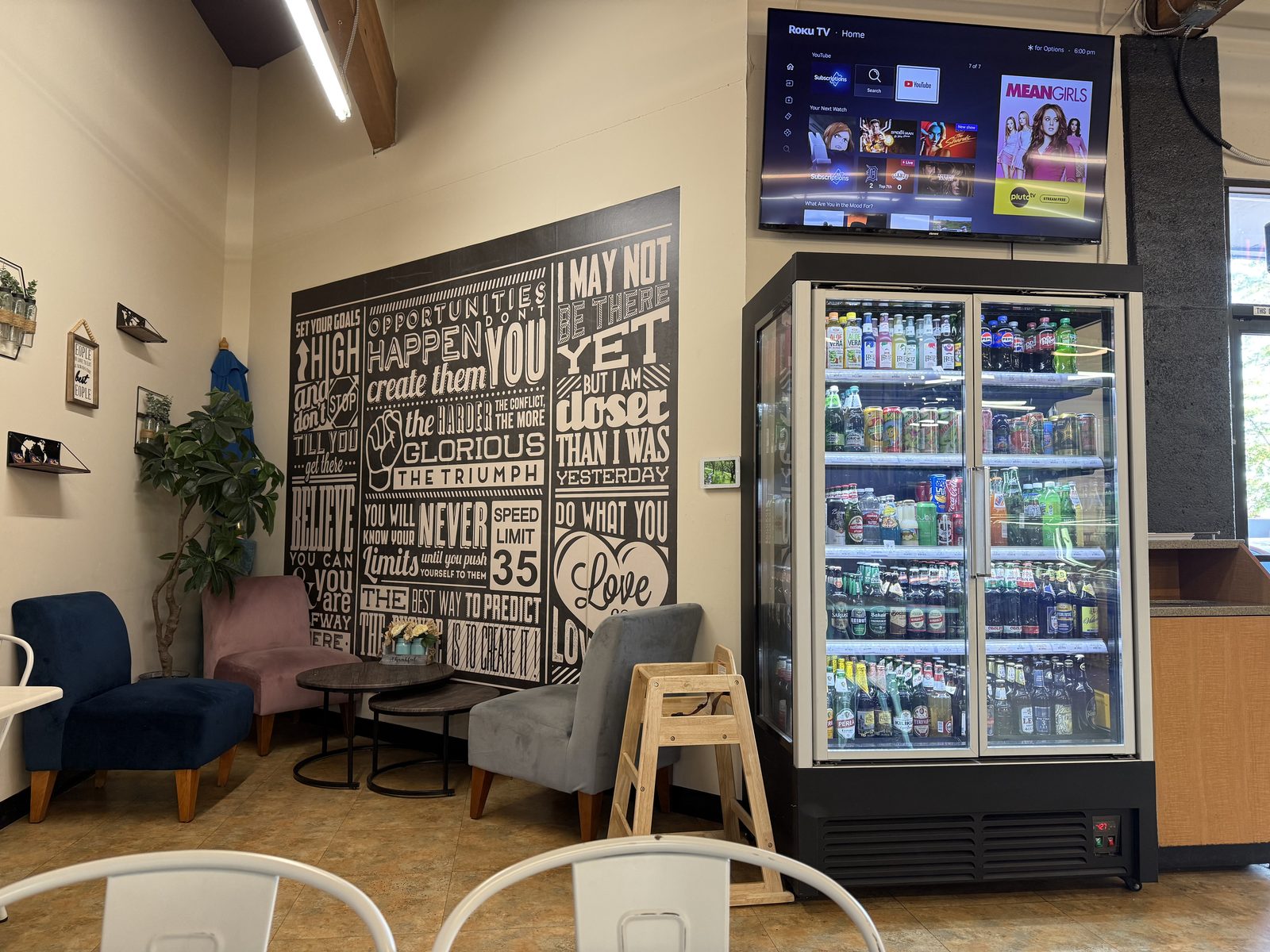 A small seating nook inside Anoush Deli with blue, pink and gray velvet armchairs around low round tables, a black and white motivational-quote wall mural, a wall-mounted TV, and a glass-front cooler stocked with soda and beer