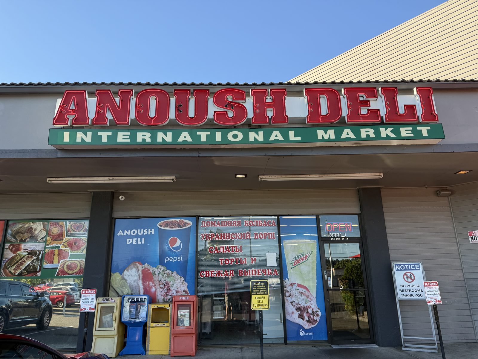 The Anoush Deli International Market storefront on NE Fourth Plain Boulevard in Vancouver, Washington, with a red neon-style ANOUSH DELI sign, a green INTERNATIONAL MARKET banner, Cyrillic window signage advertising homemade sausage and Ukrainian borscht, an OPEN sign, and No Parking Reserved for Anoush Deli Only signs beside a shared strip-mall parking lot