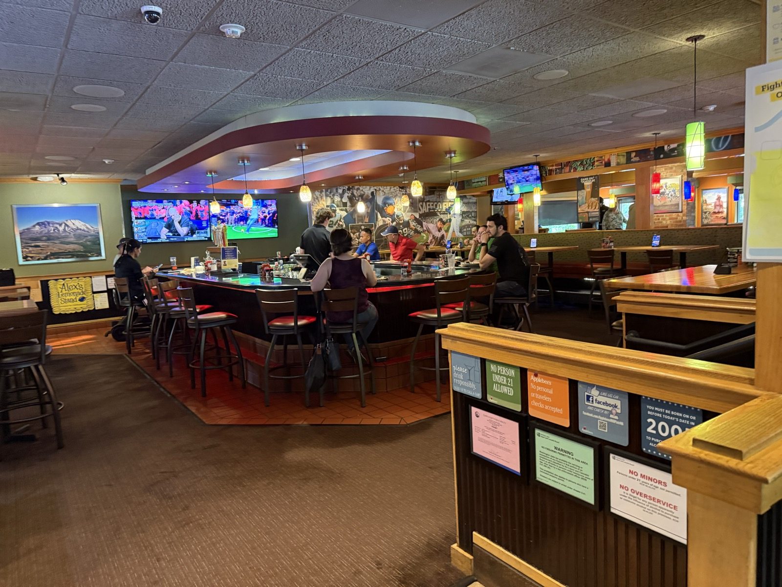 The bar at Applebee's, a rounded island bar with red padded stools, hanging pendant lights, and wall-mounted televisions showing a football game, with a handful of patrons seated around it on a weekday evening