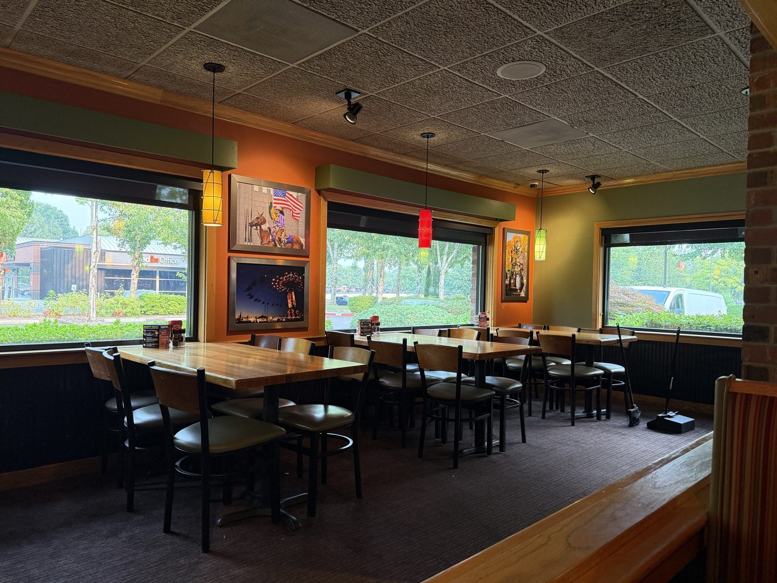 A quiet corner of the dining room with empty wood tables and chairs, framed photographs of a rodeo rider and a lit-up carnival midway hanging on an olive-green wall, and a view through the windows of the parking lot outside